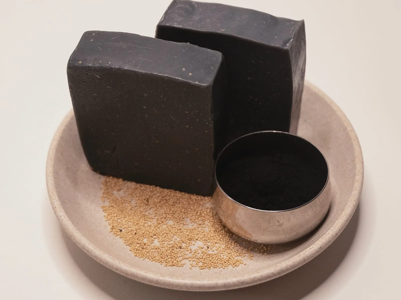 Two black activated charcoal soap bars on a ceramic dish beside a small tin of charcoal powder.
