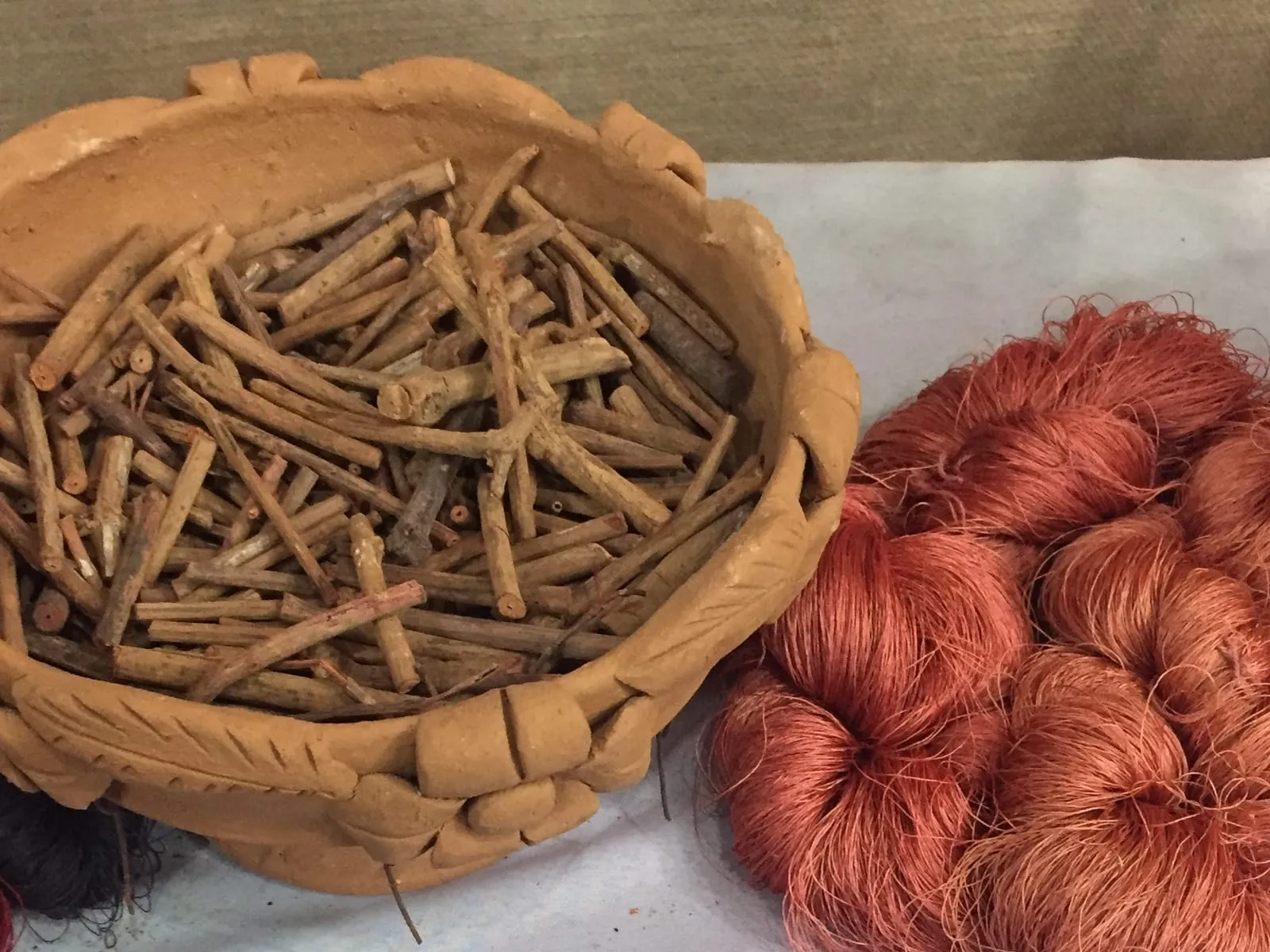 A clay basket of dried madder root sticks beside skeins of yarn dyed rust red with madder.