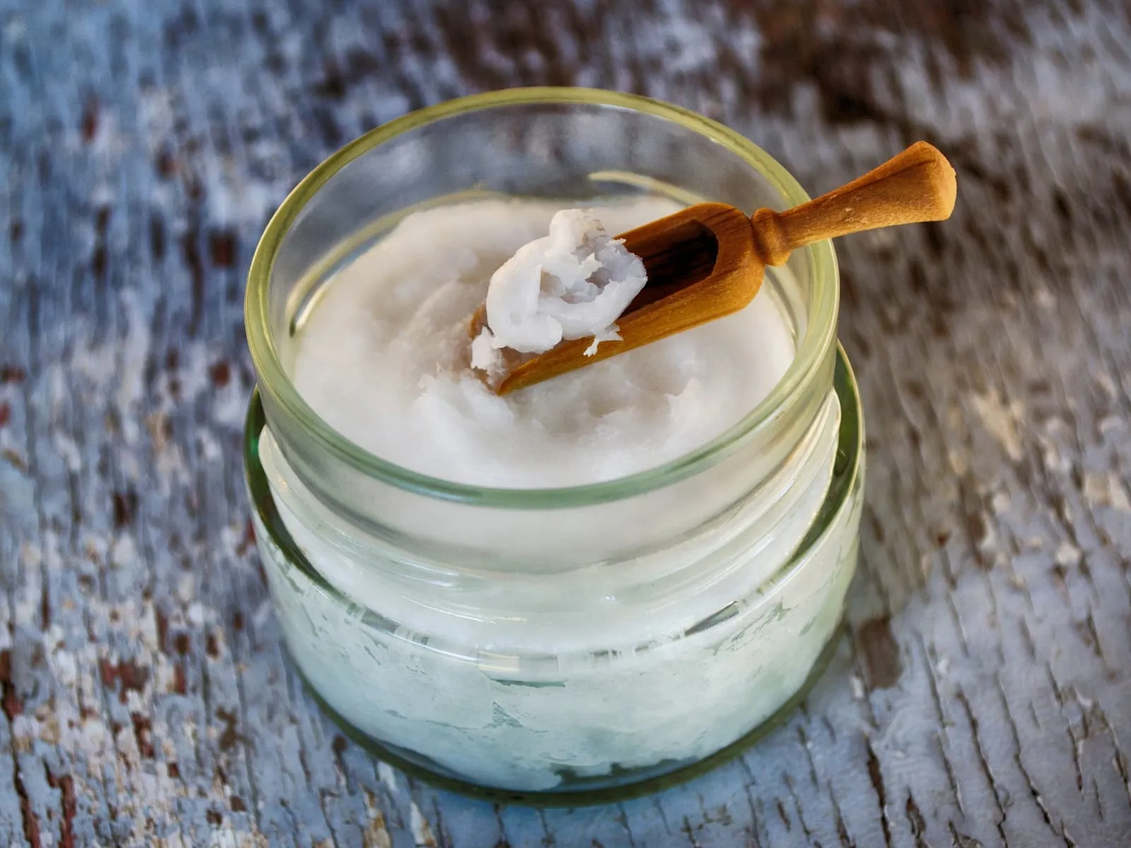 A glass jar of solid white coconut oil with a wooden scoop resting on top.